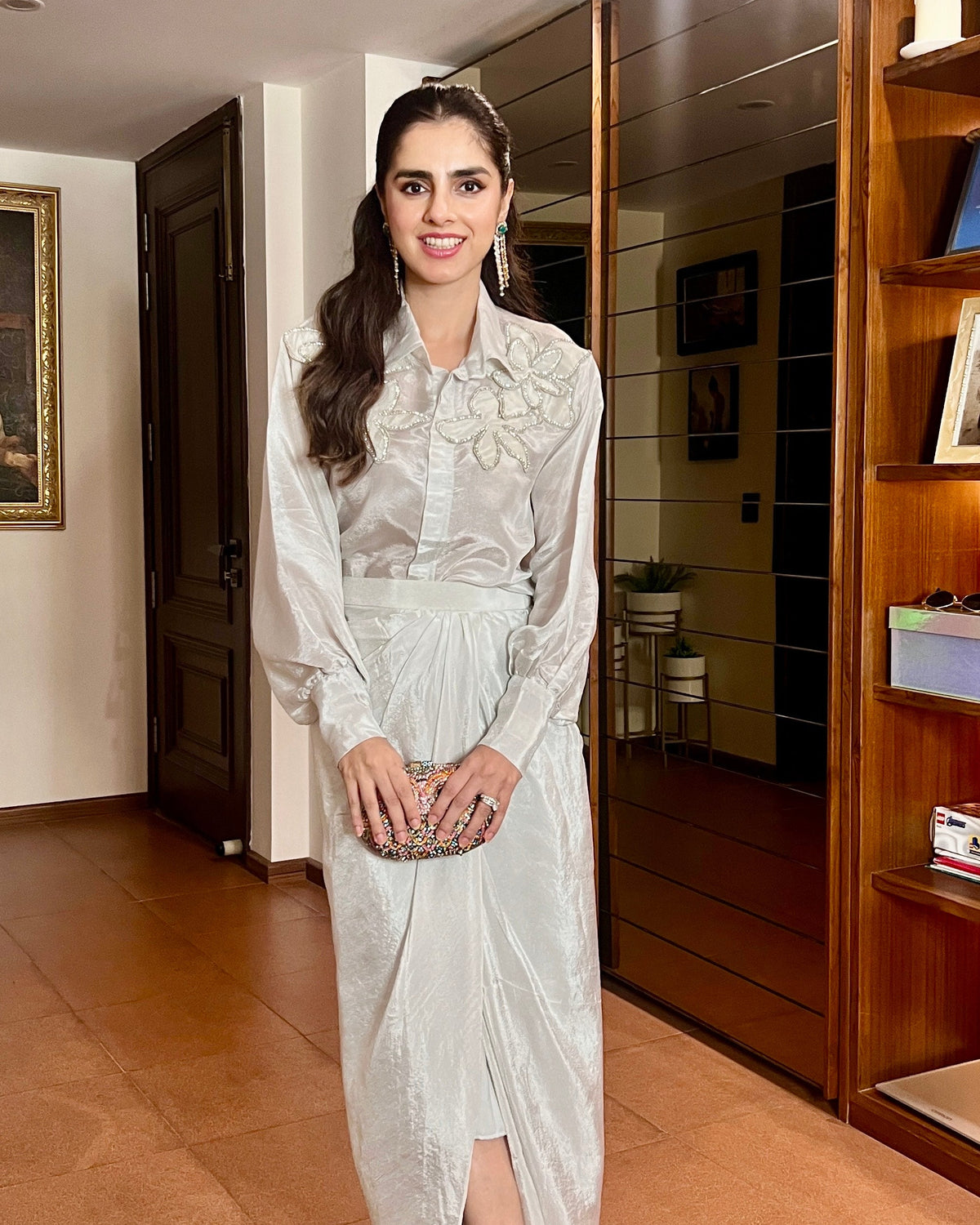 A woman wearing a silver tissue shirt with balloon sleeves and a drape skirt with flower embellishments. The outfit is formal, suited for special occasions.